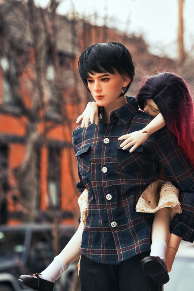 A male doll with black hair in a plaid button up shirt carrying a sleeping child doll with red and black hair on his back