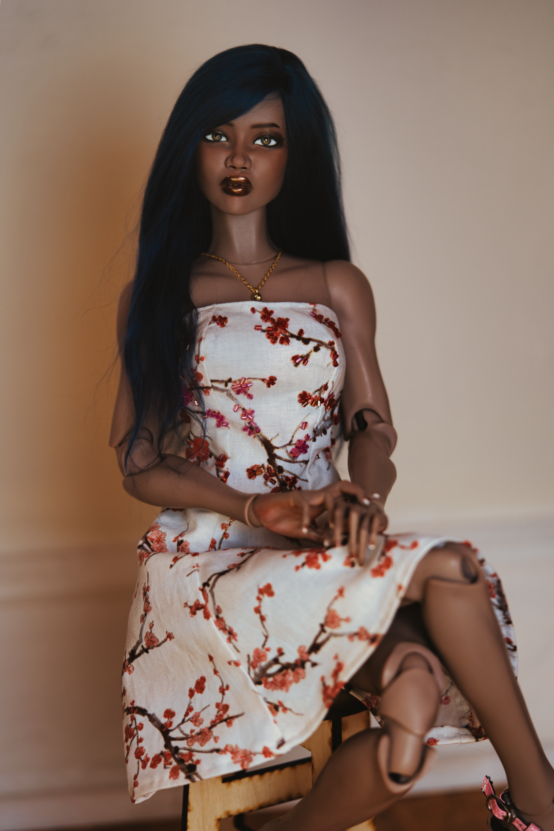 A Black female doll with long straight dark blue hair in a plum blossom dress sitting with one leg crossed over the other on a wooden stool.