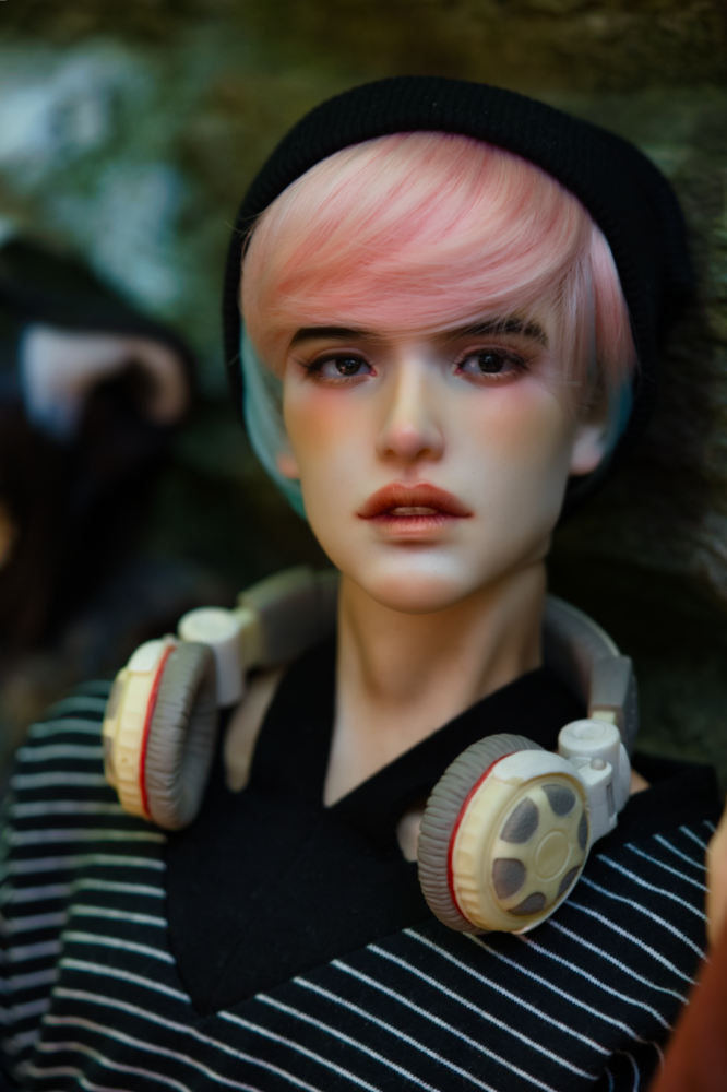 A male doll with pink and blue hair wearing a black beanie and navy shirt with thin white stripes with overear headphones around his neck.